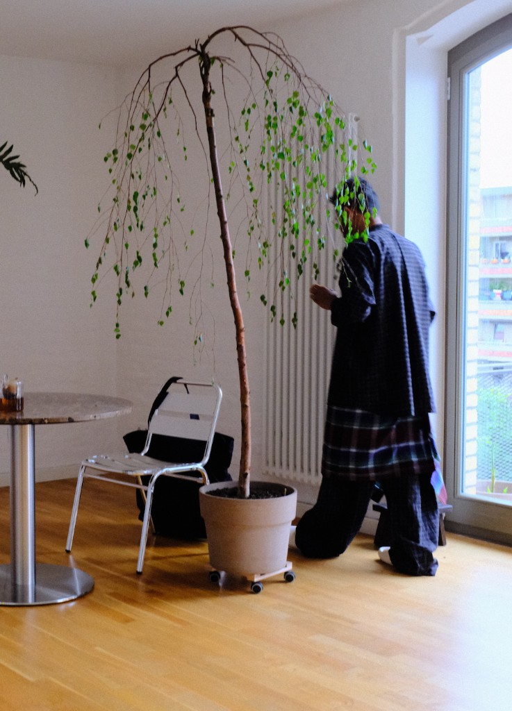 Mal standing beside a potted tree in a bright room.